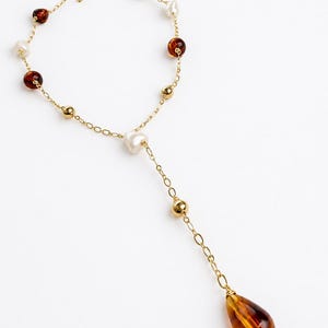 May include: A gold-tone chain necklace featuring a Y-shaped drop design. The necklace is accented with alternating round amber-colored beads, white pearl-like beads, and small gold-tone metal spheres, ending in a large teardrop-shaped amber pendant.