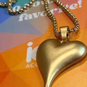 May include: Gold-colored necklace with a heart-shaped pendant. The pendant has a smooth, matte finish and a teardrop-shaped bottom. The chain is a textured, rounded design. The background includes text that says "favorite!"