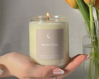 Op de afbeelding: Een brandende kaars in een heldere glazen pot met een label met de tekst "sunrise". Het label bevat de geurbeschrijving: "ORANGE, LEMON & LIME Ethereal Aroma | 8oz". Oranje en gele tulpen in een vaas.