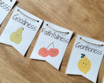 Puede incluir: Una guirnalda decorativa compuesta por banderines rectangulares blancos con base puntiaguda, unidos por un cordel marrón. Cada banderín muestra una ilustración de fruta con cara sonriente y texto: Goodness con una pera, Faithfulness con cerezas y Gentleness con un limón.