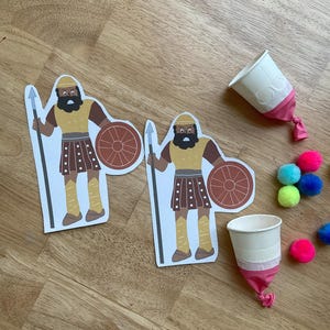 May include: Two paper cutouts of a warrior with a spear and shield, alongside two white paper cups with pink balloon bottoms and colorful pom-poms. The warrior has a brown beard, yellow helmet, and brown skirt.