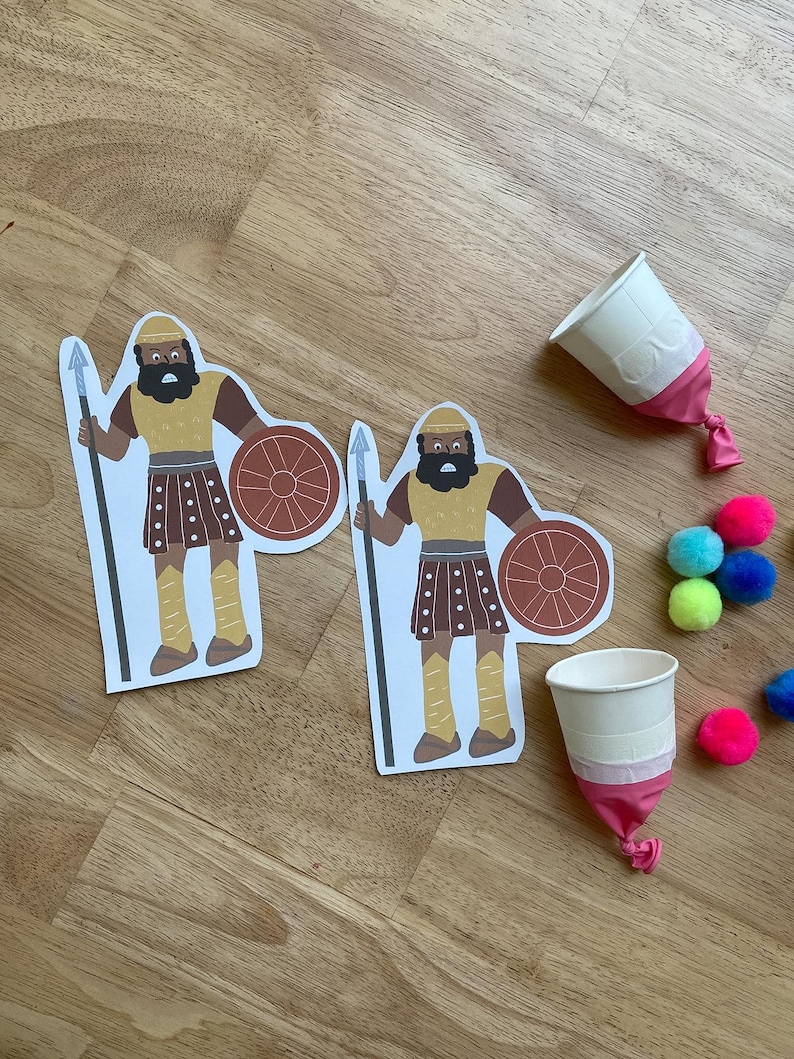 May include: Two paper cutouts of a warrior with a spear and shield, alongside two white paper cups with pink balloon bottoms and colorful pom-poms. The warrior has a brown beard, yellow helmet, and brown skirt.