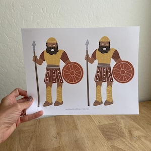 May include: A print featuring two cartoon illustrations of warriors. Each figure is depicted with a spear, shield, and wearing a yellow helmet and tunic, brown sleeves, and brown skirt. The shield is a reddish-brown color. The website address "seedsandscribbles.etsy.com" is printed at the bottom.