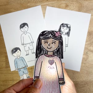 May include: A paper cutout of a person with long dark hair wearing a pink dress with a heart graphic on the chest, illuminated from behind. In the background, two white sheets of paper feature black and white line drawings of people.