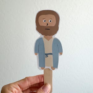 May include: A paper cutout figure of a person with a brown beard and hair, wearing a light blue robe over a beige tunic, attached to a wooden craft stick. The figure has a simple illustrated face with black eyes, eyebrows, and a straight mouth.