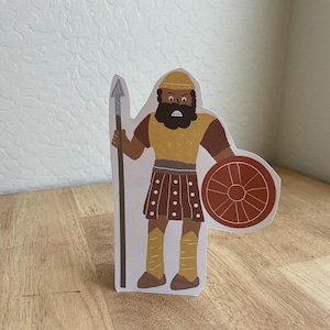 Puede incluir: Ilustración de dibujos animados de una figura de guerrero. La figura está representada en tonos marrones, beige y amarillos, sosteniendo una lanza y un escudo redondo. El guerrero lleva un casco, una armadura y una falda. Fondo de madera clara.