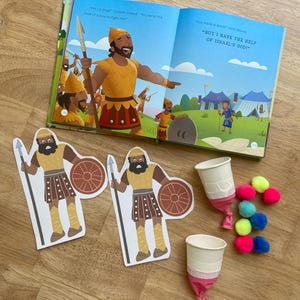 May include: An open children's book with illustrations and text, alongside cutouts of a warrior with a spear and shield, two paper cups, and colorful pom-poms. The book's text includes the phrases "Am I a dog?" and "But I have the help of Israel's God!"