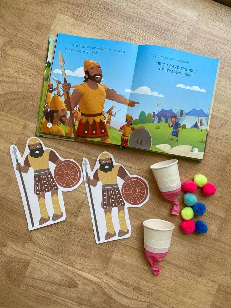 May include: An open children's book with illustrations and text, alongside cutouts of a warrior with a spear and shield, two paper cups, and colorful pom-poms. The book's text includes the phrases "Am I a dog?" and "But I have the help of Israel's God!"