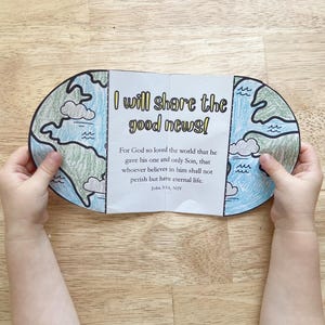 Könnte beinhalten: Ein handgefertigtes, gefaltetes Papierhandwerk, das eine Weltkarte mit blauen Ozeanen, grünen Kontinenten und Wolken darstellt. Der Text "I will share the good news!" ist zu sehen, zusammen mit einem Bibelvers. Das Handwerk wird von kleinen Händen offen gehalten.