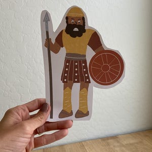 May include: A paper cutout of a warrior figure. The illustration features a figure with a yellow helmet, tunic, and leggings, brown arms and skirt, and a black beard. The warrior holds a spear and a round shield.