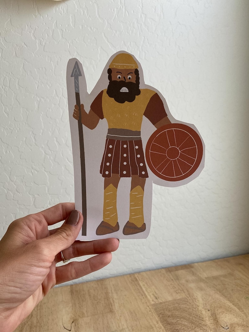 May include: A paper cutout of a warrior figure. The illustration features a figure with a yellow helmet, tunic, and leggings, brown arms and skirt, and a black beard. The warrior holds a spear and a round shield.
