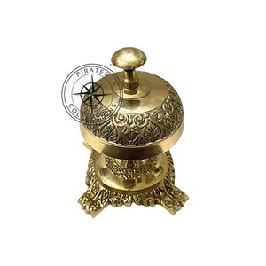 May include: A gold-colored brass bell with an ornate design and a round top. The bell sits on a three-legged base with a decorative design.