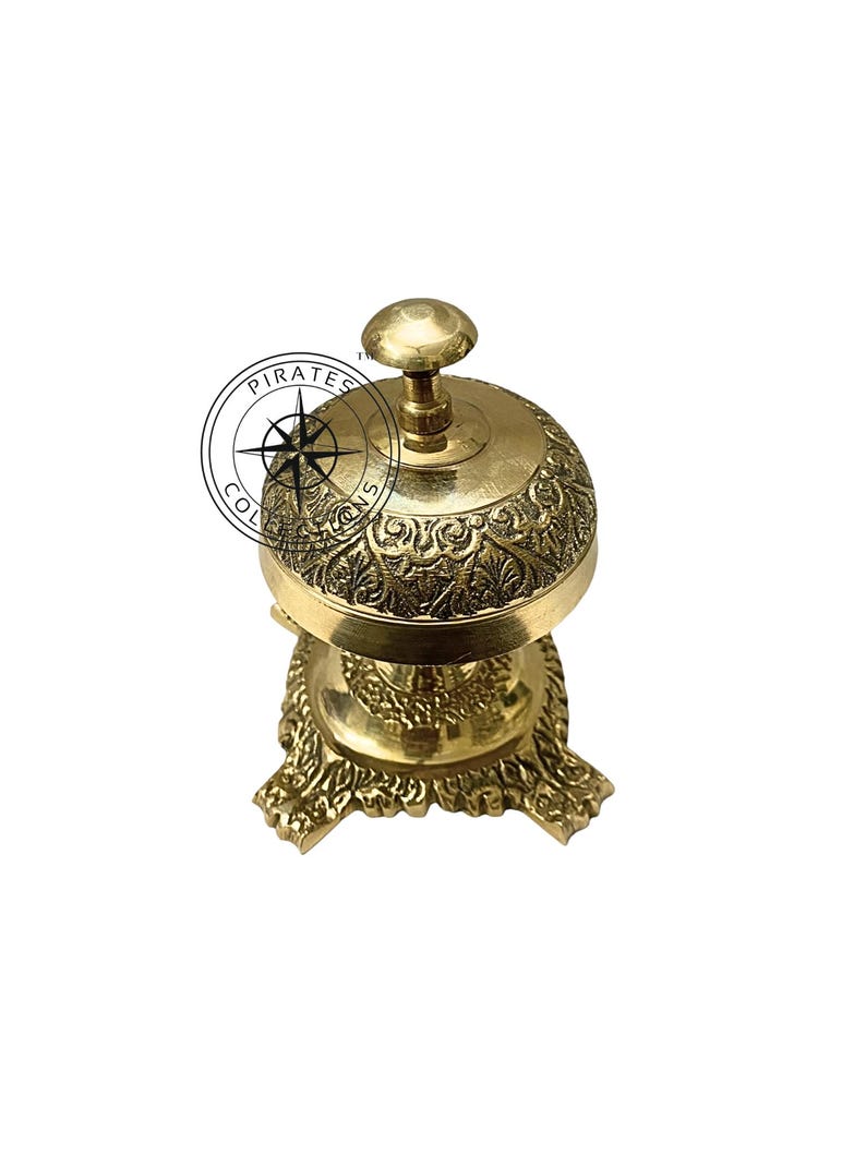 May include: A gold-colored brass bell with an ornate design and a round top. The bell sits on a three-legged base with a decorative design.