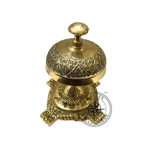 May include: A gold-colored brass bell with an ornate design on the top and base. The bell is on a stand with a decorative base.