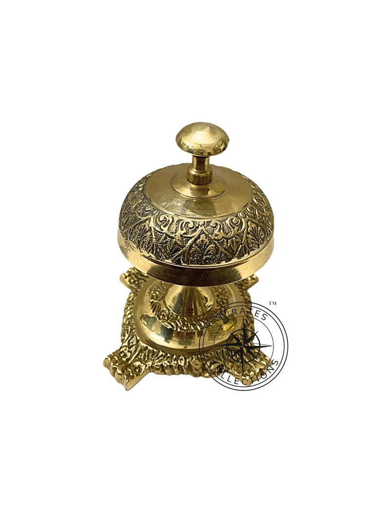 May include: A gold-colored brass bell with an ornate design on the top and base. The bell is on a stand with a decorative base.