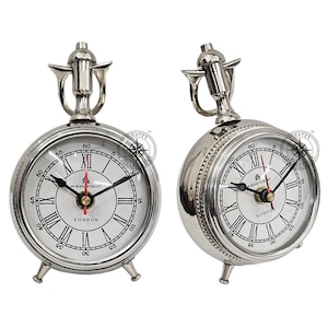 May include: Two silver-colored, vintage-style clocks with white faces, black Roman numerals, and black hands. The clocks have a decorative top and three small feet. The clock face reads "49 Bond Street London".