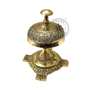 May include: A gold-toned brass service bell with an ornate design. The bell has a round top with a knob and a base with four decorative feet. The bell is engraved with a floral pattern.