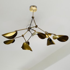 Op de afbeelding: Een moderne kroonluchter in messingkleur met bladvormige kappen. De armatuur heeft meerdere armen die uitgaan van een centraal punt, elk met een gebogen, metalen blad. Een geometrisch en elegant ontwerp voor binnenverlichting.