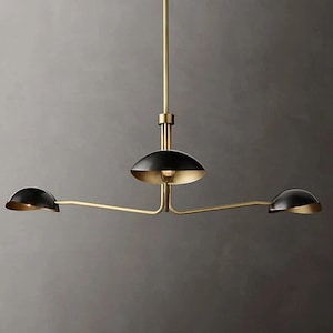 Op de afbeelding: Een moderne kroonluchter met drie lampen, een messing afwerking en zwarte kappen. De armatuur heeft een centrale stang met twee armen die naar buiten uitsteken, elk met een zwarte, halfronde kap met een gouden binnenkant.