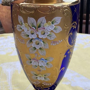 Puede incluir: Un jarrón de cristal azul cobalto con un adorno dorado y motivos florales pintados a mano en blanco y tonos pastel. El jarrón tiene un cuello acampanado, un cuerpo cónico y una base circular, decorados con intrincados detalles dorados.