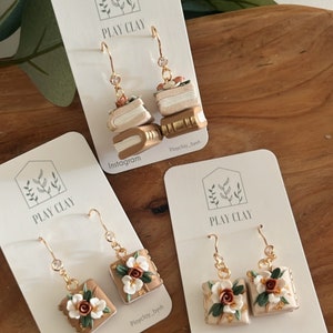 Puede incluir: Pendientes colgantes dorados con diseños detallados de arcilla polimérica. Un par presenta pilas de libros en miniatura, el otro, cuadrados florales. Cada pendiente tiene una pequeña gema y se muestra en una tarjeta blanca con el logotipo "PLAY CLAY" y el nombre de Instagram.