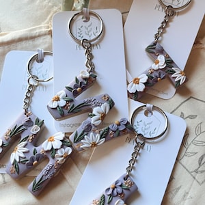 May include: Keychains with the letter "T" are displayed, each adorned with white and purple floral designs. The keychains are attached to silver chains and keyrings, presented on white cards. The background includes a beige bag and greenery.
