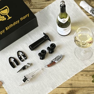 May include: A black gift box with gold text that reads "Happy Birthday Harry" and a wine glass and grape design. The image also shows a wine bottle, a glass of wine, and various wine accessories, including a corkscrew and bottle stopper.