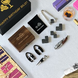 Op de afbeelding: Een assortiment van barartikelen en geschenken, waaronder een zwarte doos met gouden tekst "HAPPY BIRTHAY BILLY!", een houten doos met whisky stenen, een fles wijn en ingepakte cadeaus. Andere items zijn een kurkentrekker, flesstop en ijstang.