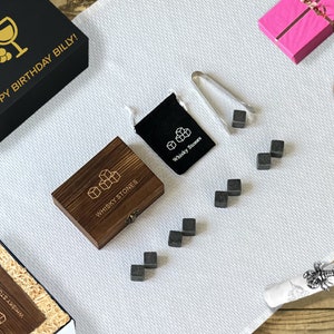 Op de afbeelding: Een verzameling whisky stenen en accessoires, waaronder een houten doos, een zwarte geschenkdoos met gouden tekst "HAPPY BIRTHDAY BILLY!" en een roze geschenkdoos. De whisky stenen zijn gerangschikt op een witte doek.