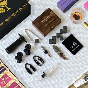 May include: A collection of bar and wine accessories, including a black gift box with gold text reading "HAPPY BIRTHDAY BILLY!", a bottle of wine, whisky stones, and various bottle stoppers and openers. The items are arranged on a white textured surface.