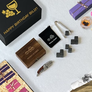 May include: A flat lay of a gift set featuring a black box with gold text reading "HAPPY BIRTHDAY BILLY!". The set includes a wooden box of whiskey stones, a bottle of wine, and wrapped gifts in purple and pink.