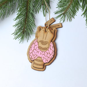 May include: A wooden Christmas ornament shaped like a thyroid gland. The ornament is painted pink and has a brown wooden tag with the text "YEAR KRISTIN" on it.