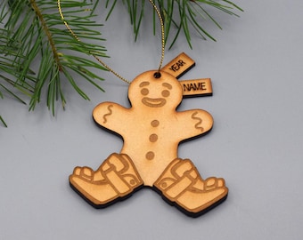 May include: A light brown wooden gingerbread person ornament with a gold hanging string. The figure features etched facial details, three buttons, and medical leg braces. Two small rectangular wooden tags are attached, labeled YEAR and NAME.