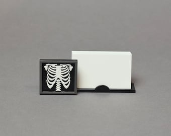 May include: A black and white ribcage design on a small black square card holder. The card holder is next to a white card holder with a black base.