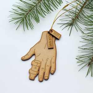May include: Wooden Christmas ornament shaped like a hand with a broken finger in a cast. The ornament has a tag that says "YOUR NAME" and another tag that says "2023".