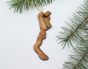 May include: Wooden Christmas ornament shaped like a state with a tag that says "YEAR" and "OUR NAME". The ornament is hanging from a gold string and is surrounded by pine branches.