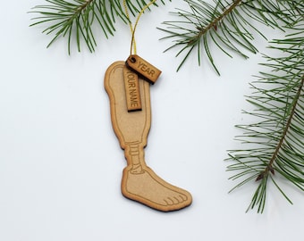 May include: Wooden Christmas ornament shaped like a prosthetic leg with two tags attached. The tags are labeled "YOUR NAME" and "YEAR".