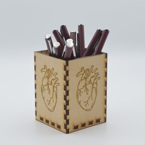 May include: A wooden pencil holder with a heart design. The holder is filled with red pens.