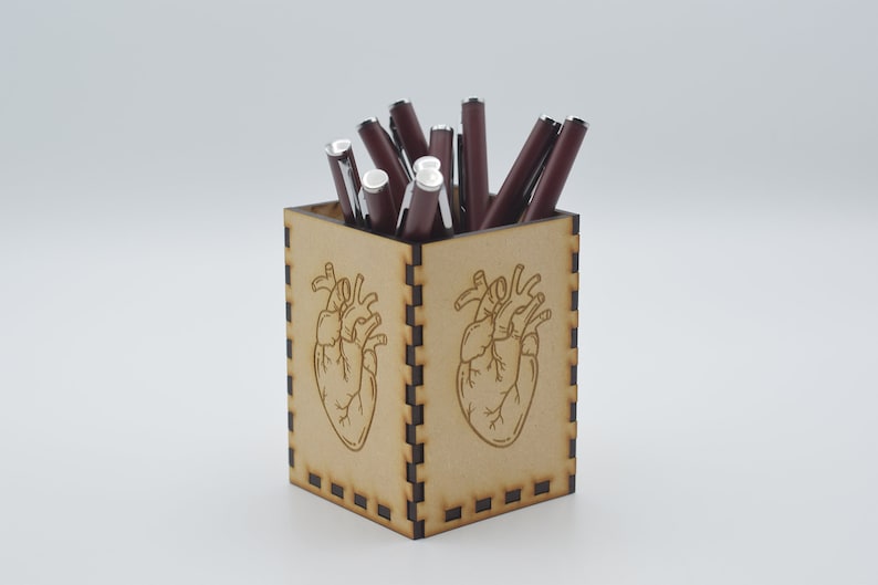 May include: A wooden pencil holder with a heart design. The holder is filled with red pens.