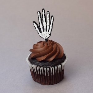 May include: A chocolate cupcake with chocolate frosting topped with a black skeleton hand cupcake topper.