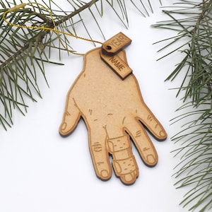 May include: Wooden Christmas ornament shaped like a hand with a bandaged finger. The ornament has two tags attached to it, one that says "YEAR" and one that says "NAME".