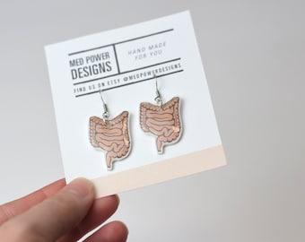 Pode incluir: Um par de brincos pendentes com uma ilustração anatômica detalhada dos intestinos humanos. Os brincos são feitos de acrílico transparente com um esquema de cores marrom e bege. Os brincos são feitos à mão e o cartão diz "Med Power Designs" e "Find us on Etsy @medpowerdesigns".