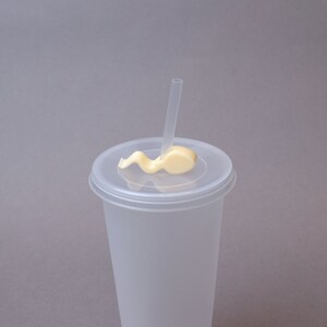 Puede incluir: Un vaso de pl&aacute;stico transparente con tapa y una pajita. La tapa tiene un remolino de glaseado blanco en la parte superior.