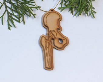 May include: A wooden ornament shaped like a human hip bone and femur. The ornament is light brown and has a gold string attached to it.