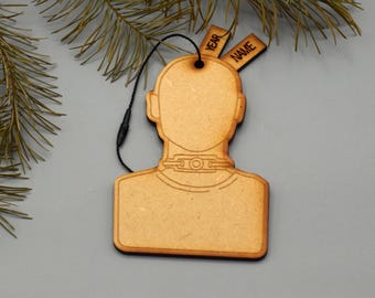 May include: A wooden ornament in the shape of a person's head and shoulders. The ornament has a black string for hanging and two small tags that read "YEAR" and "NAME". The ornament is made of light brown wood and is set against a gray background.