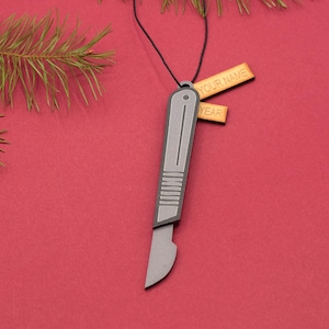 May include: A silver-colored metal ornament shaped like a scalpel with a wooden tag that says "YOUR NAME" and another tag that says "YEAR".