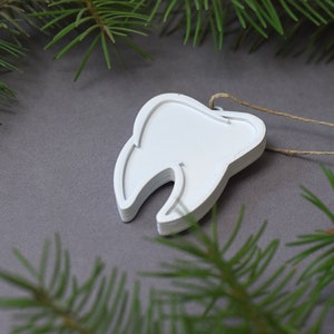 May include: White wooden tooth ornament with a string attached. The ornament is on a gray background with green pine branches.