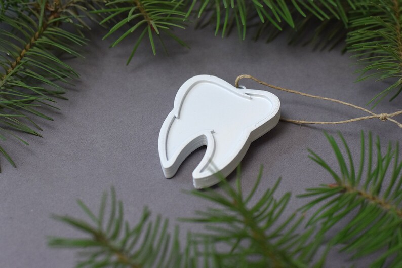 May include: White wooden tooth ornament with a string attached. The ornament is on a gray background with green pine branches.