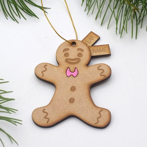 May include: A wooden gingerbread man Christmas ornament with a pink bow tie and the word "NAME" and "YEAR" on separate tags. The ornament is hanging from a gold string. Green pine needles are in the background.