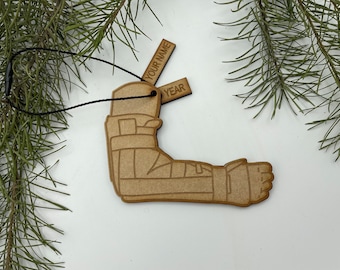 May include: Wooden Christmas ornament shaped like a broken arm in a cast. The ornament has a string for hanging and two blank tags that say "YOUR NAME" and "YEAR".